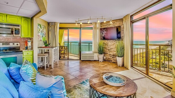 Una cocina moderna con armarios verdes, un comedor con mesa y sillas, y un balcón con vistas al atardecer.