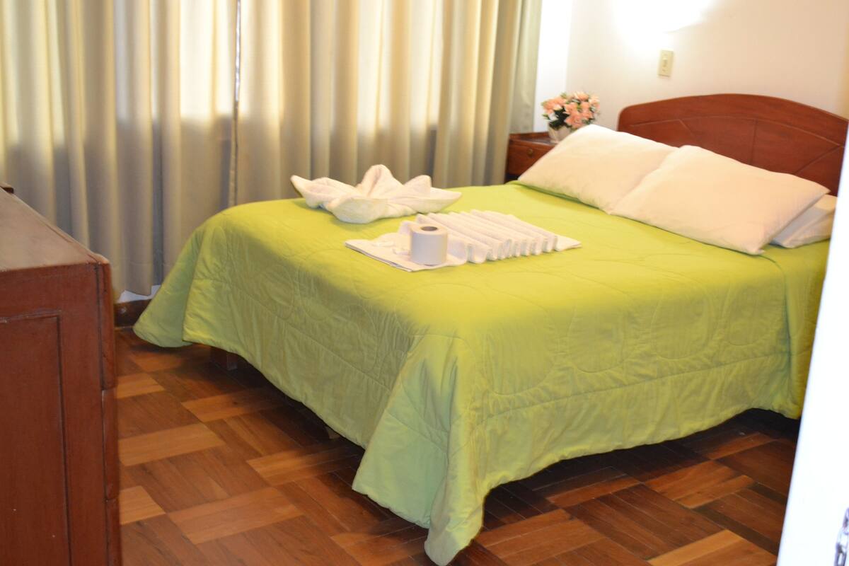 Habitación de hotel con colcha verde, suelo de madera, cabecero de madera y mesita de noche con un florero.