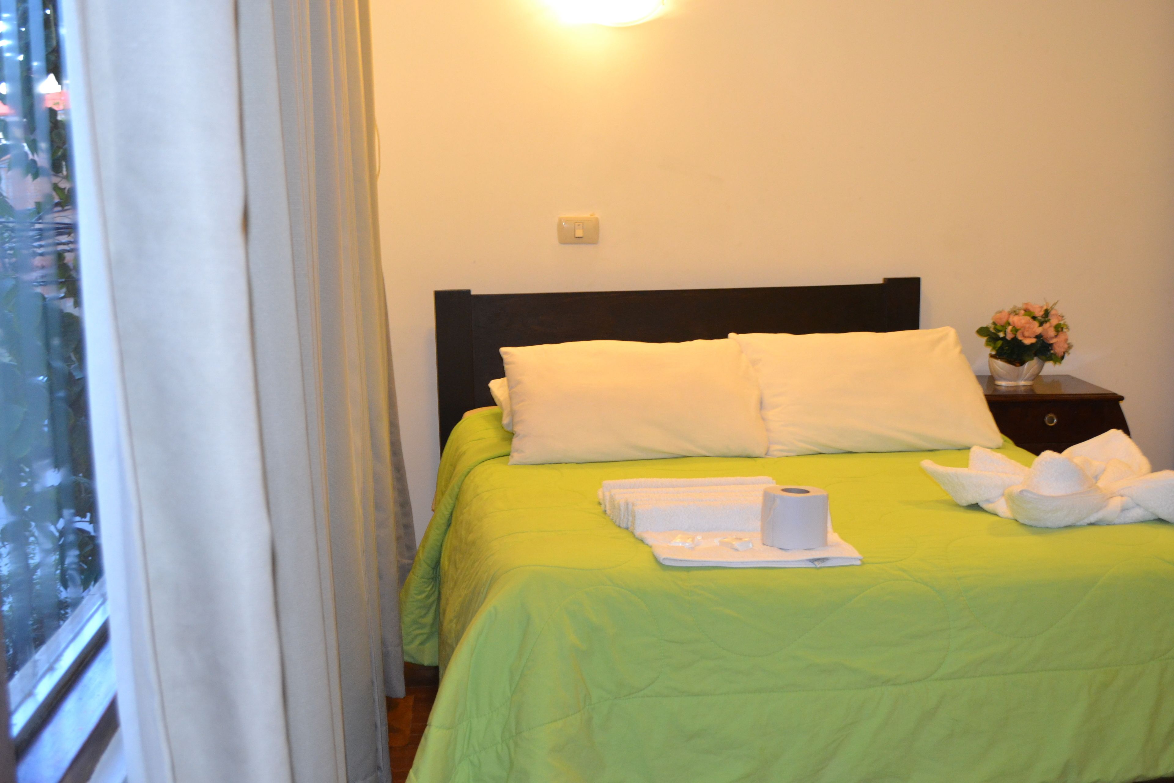 A hotel room with a bed, pillows, a nightstand with a vase, and a window with curtains.