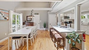 A modern kitchen with white cabinetry, a central island, and a dining area with a white table and chairs. - Shore House 003-DP Newly renovated in Dennis Port, 2 minute walk to the beach! (Dennis Port)