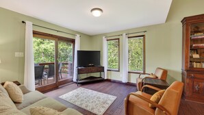 A living room with a sofa, a television on a stand, a wooden cabinet, and a view through large windows. - Lone Bear Cabin: EV, River Access, Mountain Views, Fireplace, Hot Tub (Rileyville)