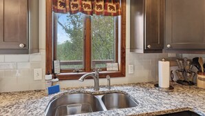 A kitchen with a double sink, wooden cabinets, a window with a plaid curtain, and a sign requesting visitors to use the door and windows. - Lone Bear Cabin: EV, River Access, Mountain Views, Fireplace, Hot Tub (Rileyville)