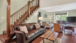 A living room with a brown leather sofa, wooden staircase, and a flat-screen TV. - Lone Bear Cabin: EV, River Access, Mountain Views, Fireplace, Hot Tub (Rileyville)