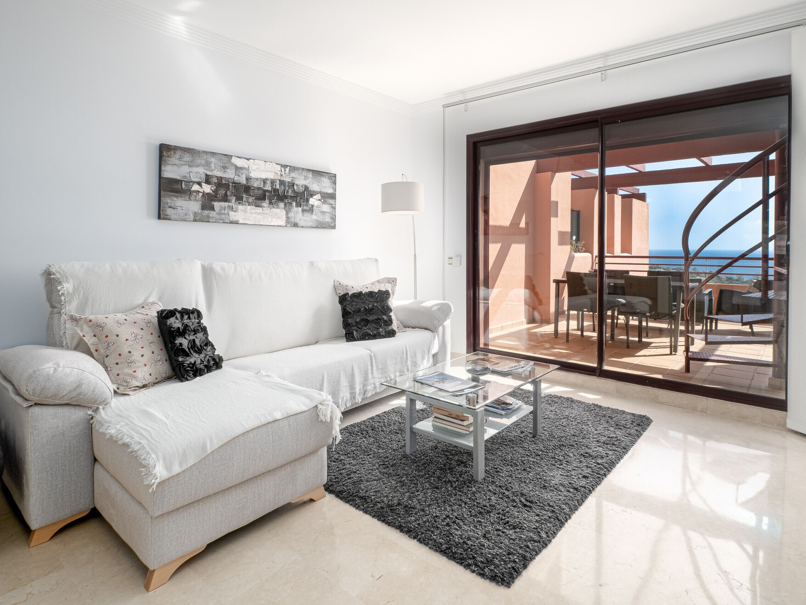A modern living room with a white sofa, a glass coffee table, and a black rug. There is a large window with a view of the sea and a balcony with outdoor furniture.