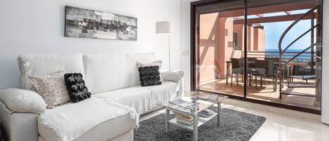 A modern living room with a white sofa, a glass coffee table, and a black rug. There is a large window with a view of the sea and a balcony with outdoor furniture.