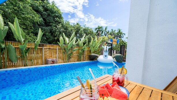 A poolside area with a wooden table, a glass of iced drink, watermelon slices, and a plate of fruit.