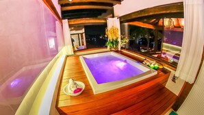Una terraza de madera con un jacuzzi, un cuenco blanco pequeño con una flor y una ventana con vista a la ciudad de noche.