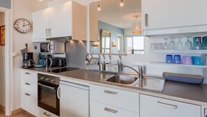 Una cocina moderna con armarios blancos, electrodomésticos de acero inoxidable y un horno empotrado. Hay un lavamanos con un grifo moderno y una cafetera en la encimera.