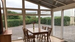 A conservatory with a wooden table and chairs, a vase with flowers, and a view of a gravel path and greenery outside.