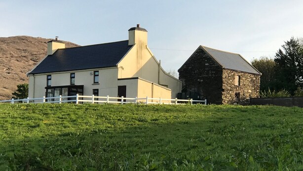 A two-story house with a white fence in front, set in a grassy field with a stone building nearby.