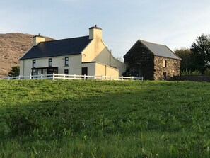 A two-story house with a white fence in front, set in a grassy field with a stone building nearby.