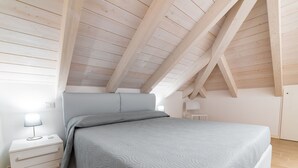 Une chambre avec un plafond en pente, un lit recouvert d’une couette grise, une table de chevet avec une lampe et une chaise.