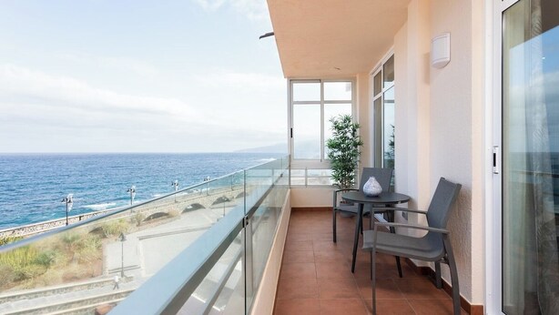 Un balcón con vistas al mar, barandilla de vidrio y una mesa con sillas.