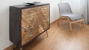 Una habitación moderna con un aparador de madera con estampado de espiga, un espejo redondo, una silla gris y un cuadro en la pared.