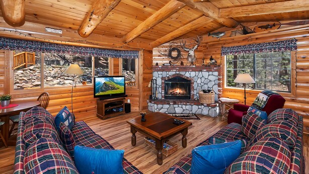 A cozy log cabin interior with a stone fireplace, a television, and a sofa with plaid cushions.