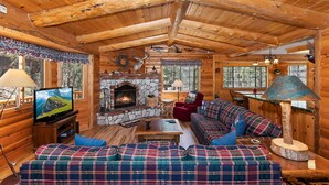 L’intérieur d’un chalet confortable, avec des murs en rondins de bois, une cheminée en pierre, des meubles à motifs écossais et un téléviseur.