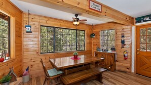 A wooden cabin interior with a dining table, benches, a ceiling fan, large windows, and a sign reading "Home Cooking".
