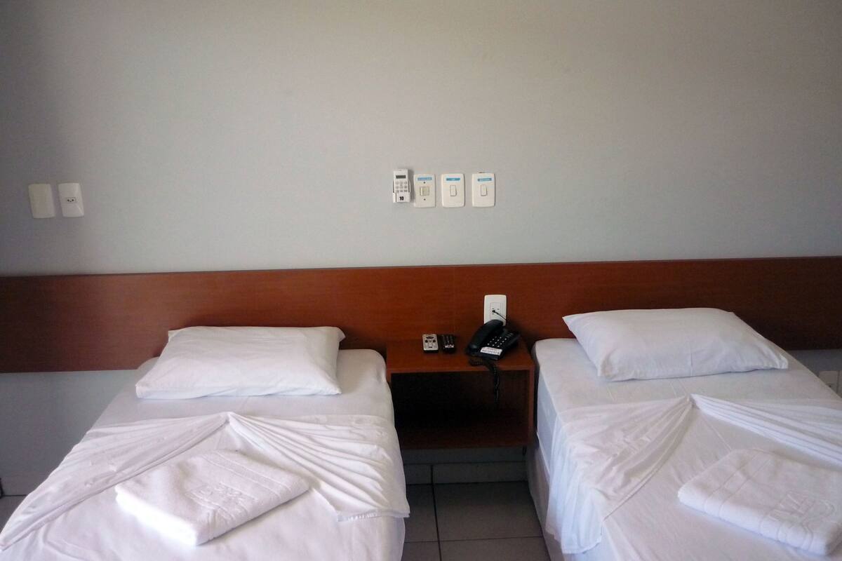A hotel room with two single beds, a wooden headboard, a bedside table with a phone and remote controls, and a wall-mounted power outlet.