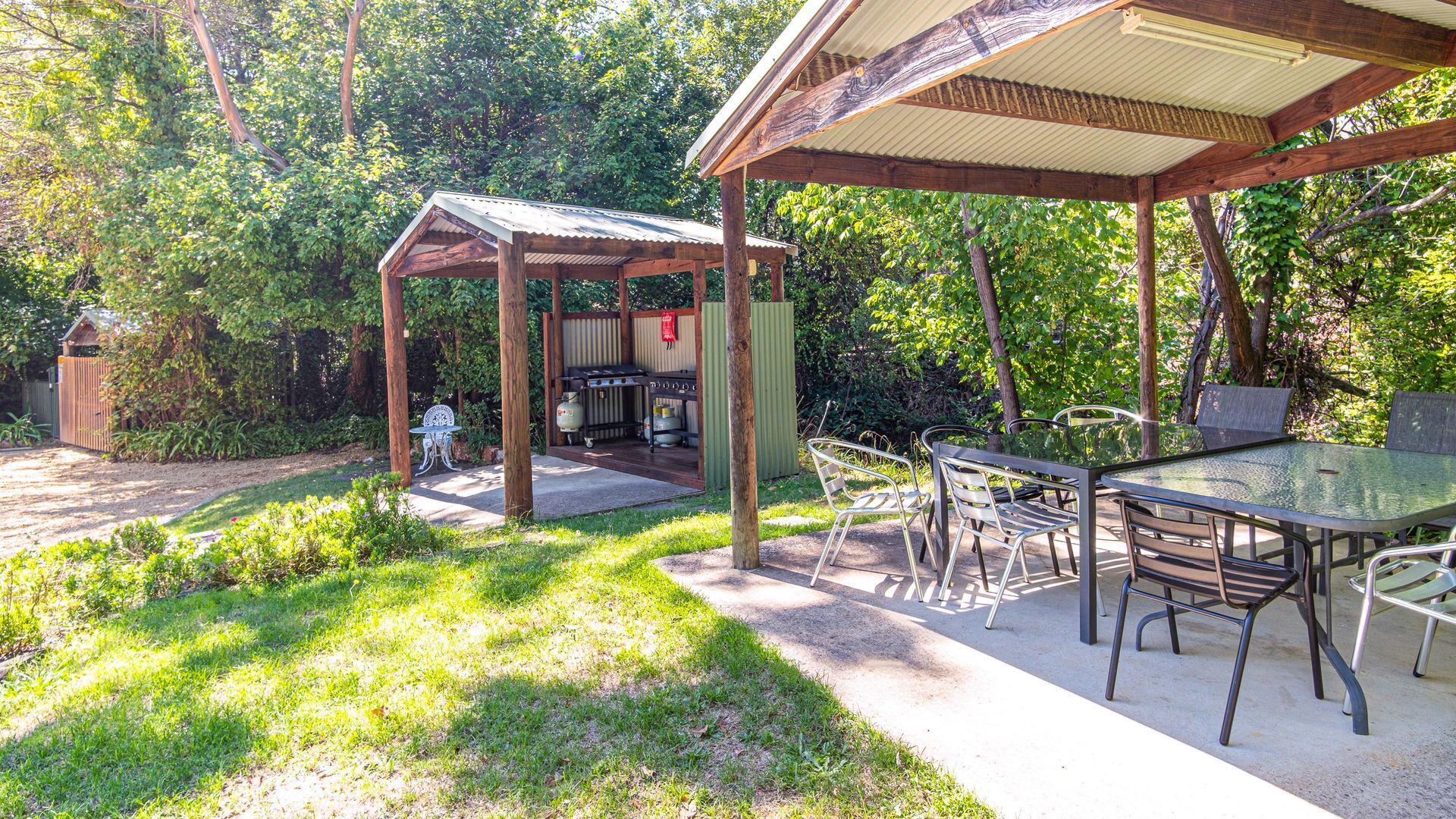 A covered outdoor area with a table and chairs, surrounded by greenery.