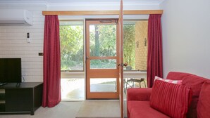 Une pièce dotée d’une porte-fenêtre coulissante donnant sur un balcon, d’un canapé rouge, d’un meuble TV noir et d’un mur en briques blanches.