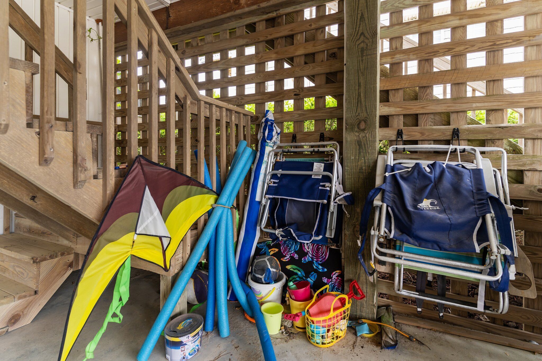 A covered outdoor area with a wooden lattice structure, a wheelchair, and various items like a kite, buckets, and hoses.