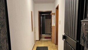 Un couloir étroit, avec un paillasson noir menant à une porte, un sol carrelé et un luminaire au plafond.