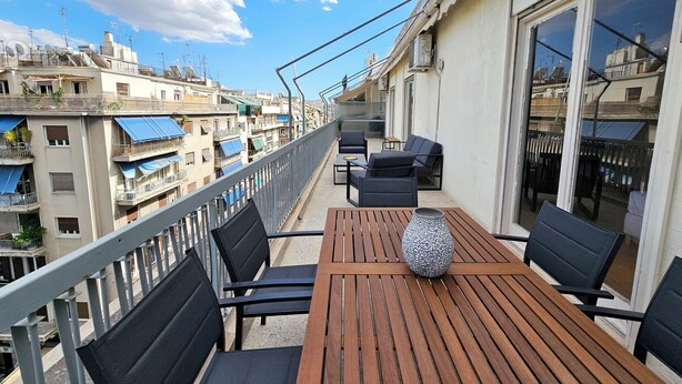 A balcony with a wooden table and black chairs, overlooking a cityscape with multiple buildings and balconies. - Victoria Capitan Apartments (Athina)