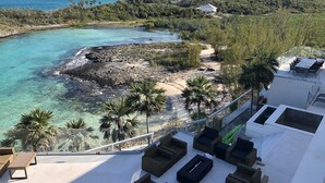Eine Dachterrasse mit Korbmöbeln, mit Blick auf eine Küstenlandschaft mit klarem türkisblauem Wasser und einem felsigen Strand.