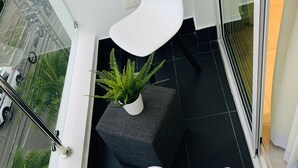 Un balcón moderno con barandilla de vidrio, dos sillas blancas, una planta en maceta y vista a la calle.