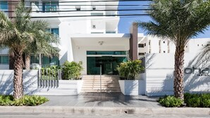 Un edificio moderno con fachada blanca, entrada de vidrio y una escalera que lleva a la entrada. Palmeras y una cerca metálica en el frente.