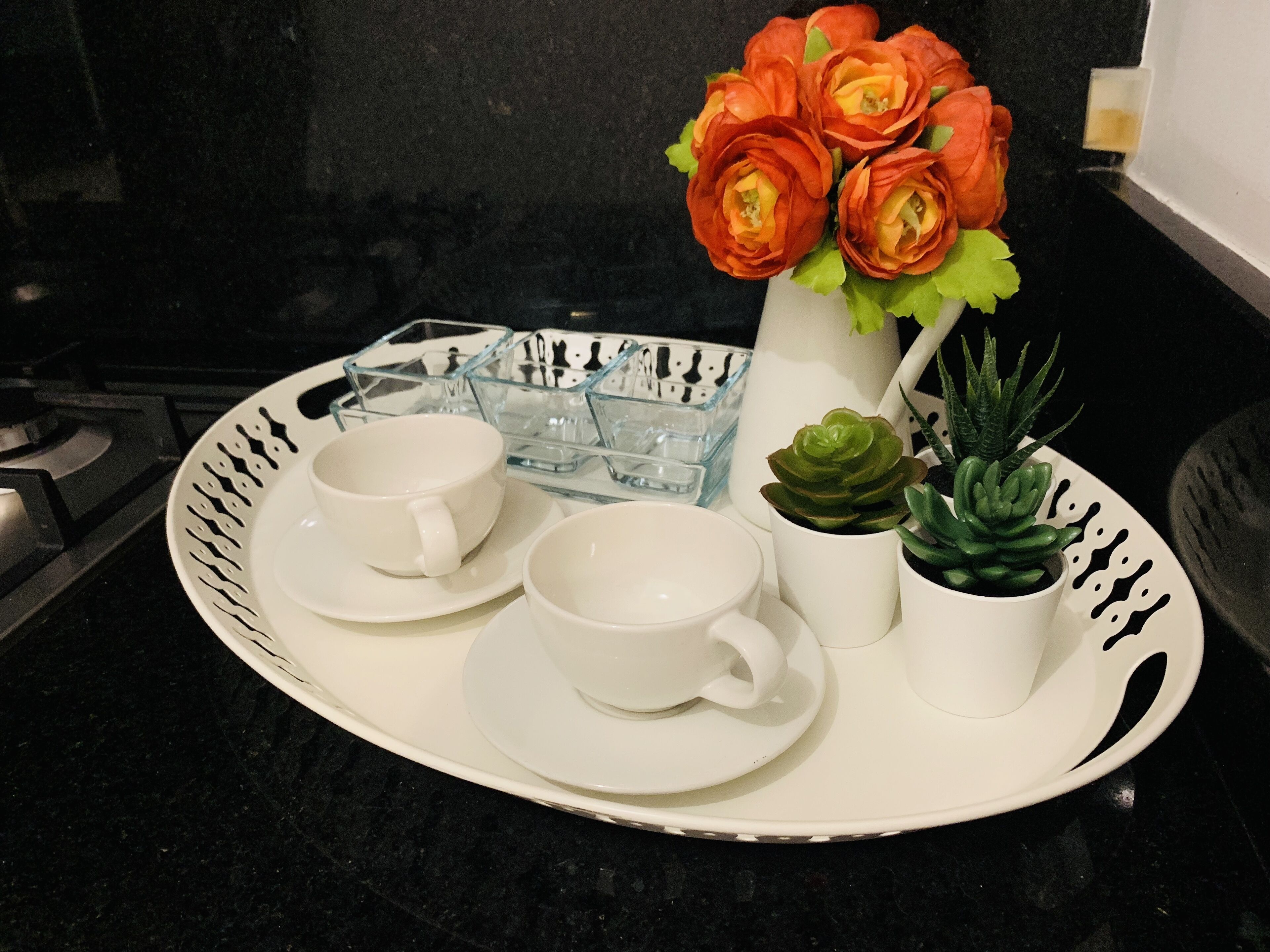 A tray with two white cups and saucers, a glass container, a vase with orange flowers, and two small potted plants.