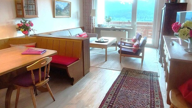 A cozy room with a wooden dining table, a sofa with a red cushion, a small table with a chair, and a view of the outdoors through a large window.
