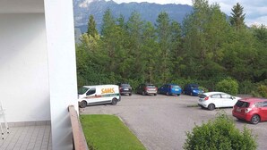 A parking area with several cars and a van, surrounded by trees and mountains in the background.
