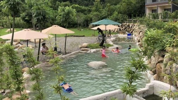 A hot spring area with people relaxing and swimming, surrounded by rocks and greenery. - Ming Yueju (Guoxing)