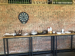 A brick wall with a dartboard and a buffet table with various food items and condiments.