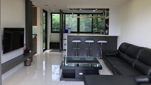 A modern living room with a black leather sofa, a glass coffee table, and a TV mounted on the wall. There is a kitchen area with a bar counter and stools, and a large window providing a view of greenery outside. - Ming Yueju (Guoxing)