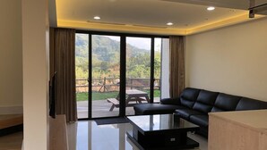 A modern living room with a black leather sofa, a glass coffee table, and a large sliding glass door leading to an outdoor patio with a wooden table and chairs. - Ming Yueju (Guoxing)
