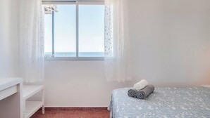 A bedroom with a bed, a window with sheer curtains, a small white shelf, and a view of the sea. - Bright 3 Bed, Sea Views Beach Apt (Benalmádena)