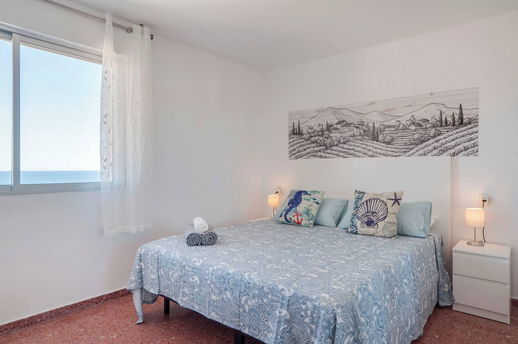Un dormitorio con cama, mesita de noche, lámpara, ventana con cortina y un cuadro de paisaje en la pared.