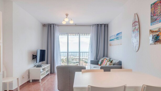 A modern living room with a TV, white furniture, a balcony with curtains, and a surfboard on the wall. - Bright 3 Bed, Sea Views Beach Apt (Benalmádena)