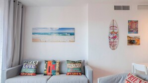 A living room with a grey sofa, a glass coffee table, and a white armchair. There are four colorful pillows on the sofa, a beach painting on the wall, and a surfboard decoration. - Bright 3 Bed, Sea Views Beach Apt (Benalmádena)
