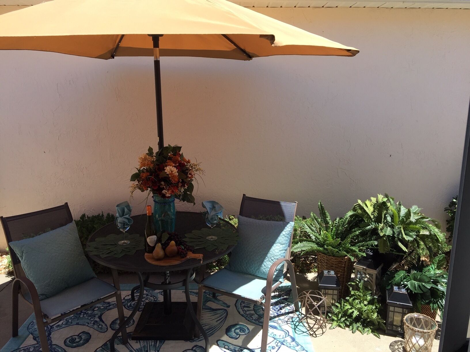A round table with a beige umbrella, two chairs with cushions, a vase with flowers, and a small table with a bottle and fruit.