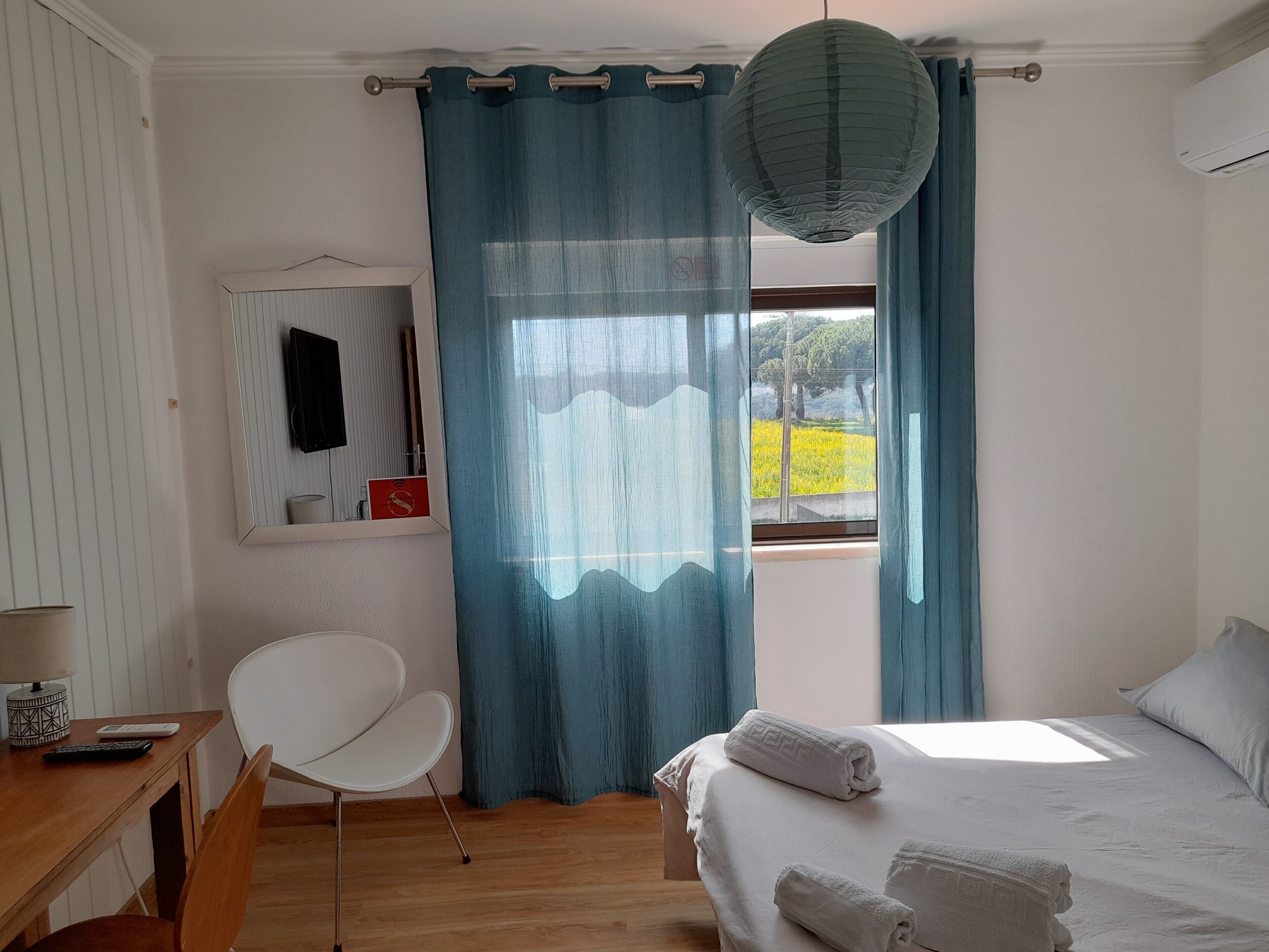 Un dormitorio con cama, una silla, un escritorio, una ventana con cortinas y una lámpara colgante.