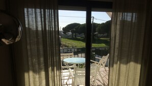 A view through a glass door with white curtains, revealing a balcony with a round table and white chairs, and a green landscape in the background. - Sublime Sun & Van, By Meco Stays (Sesimbra)