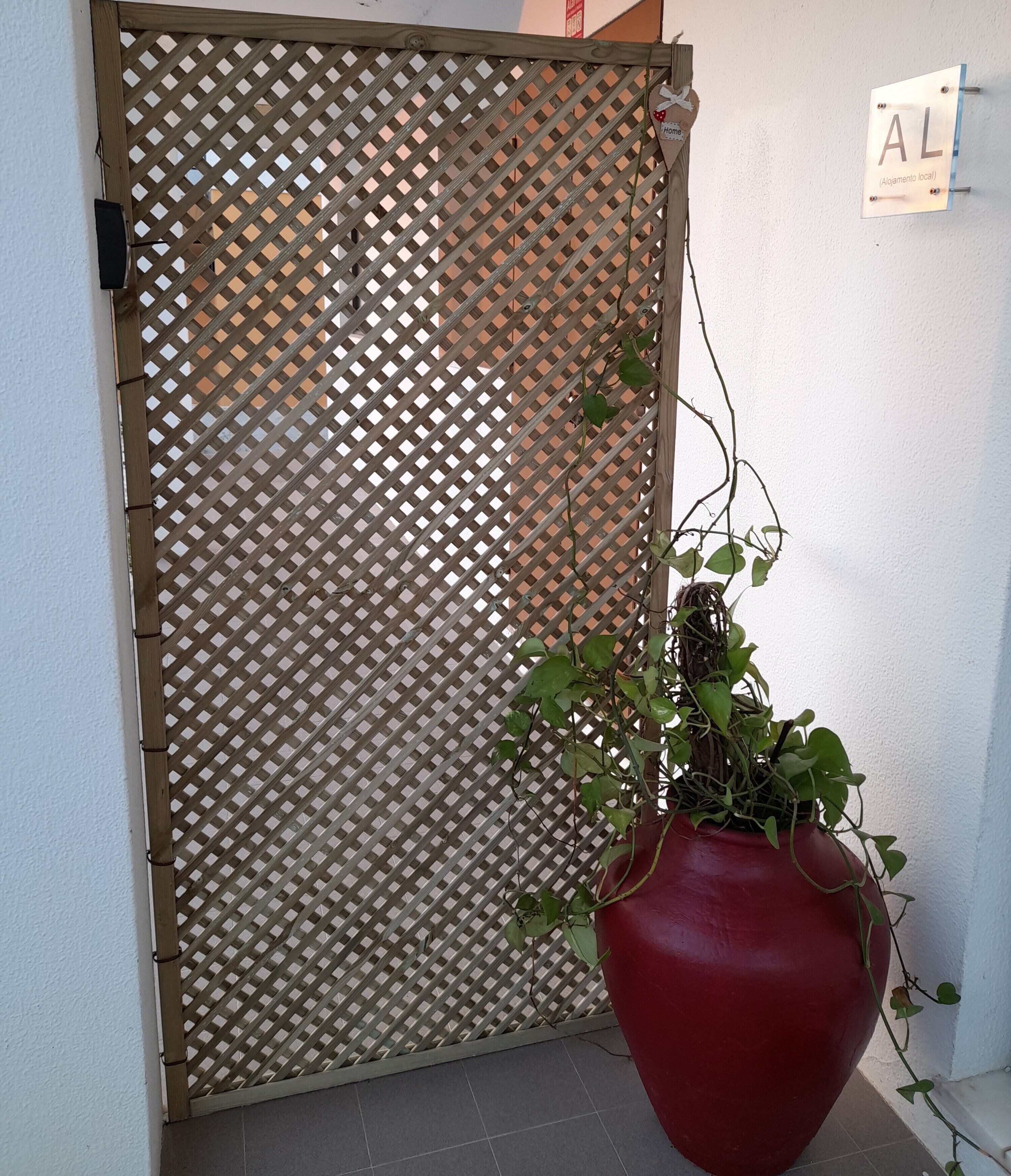 A wooden lattice gate, a red potted plant, and a sign with the letters "AL" on a white wall.
