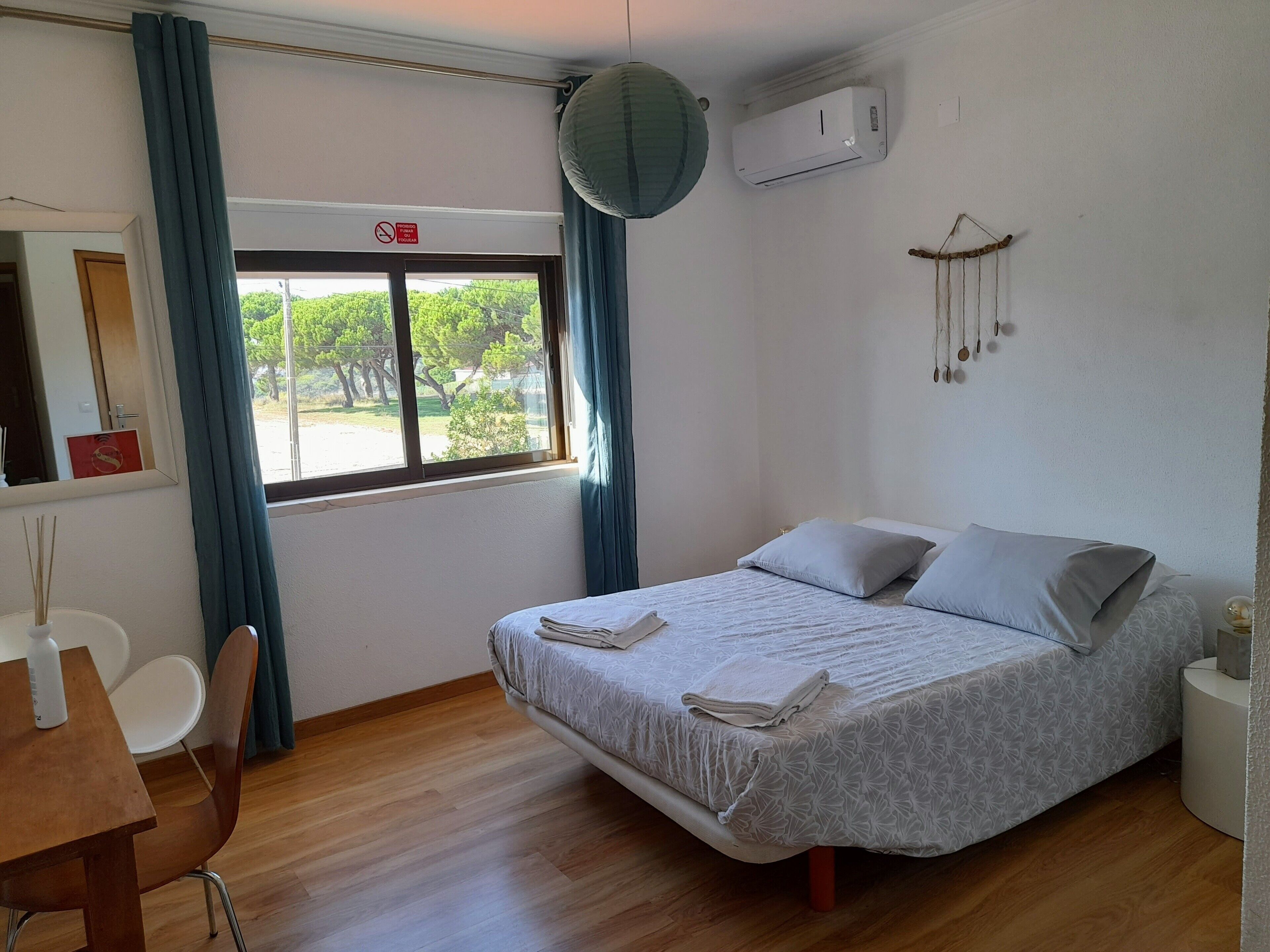 Un dormitorio con cama, escritorio, silla, ventana con cortinas, aire acondicionado y un adorno en la pared.