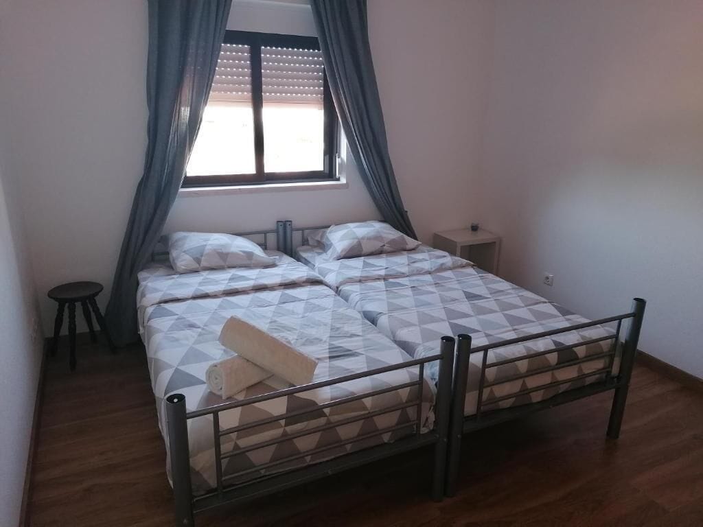 Una cama doble con estructura metálica, una mesita de noche pequeña y un ventanal con cortinas.