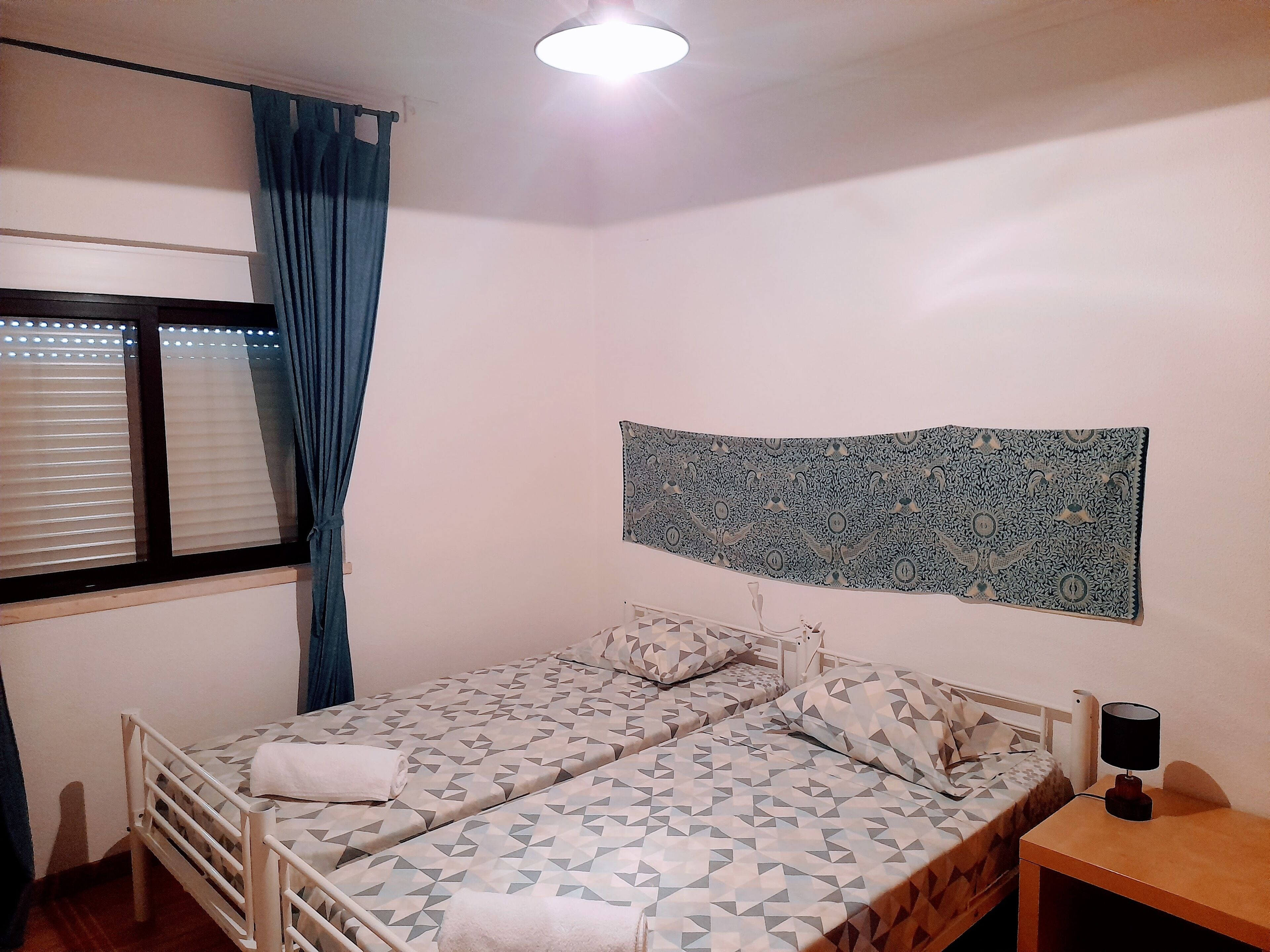 Une petite chambre propre, avec deux lits, une fenêtre avec des stores, une tapisserie murale et une lampe de chevet.