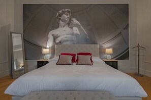 A bedroom with a large bed, two bedside tables with lamps, a large artwork on the wall, and a mirror on the left.