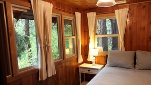 Quarto de cabana de madeira com cama, mesa de cabeceira e luminária, e janelas com cortinas.